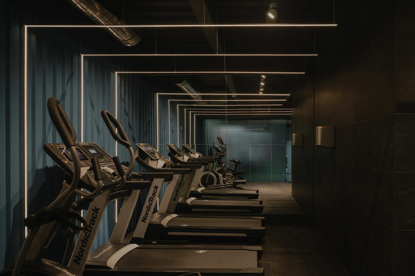 Dark gym with a row of NordicTrack treadmills, black walls, and ceiling lights creating geometric lines.