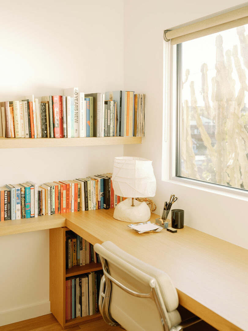 A minimalist home office with a wooden desk, chair, and bookshelves. A lamp, pens, and a view of cactus plants outside the window complete the scene.
