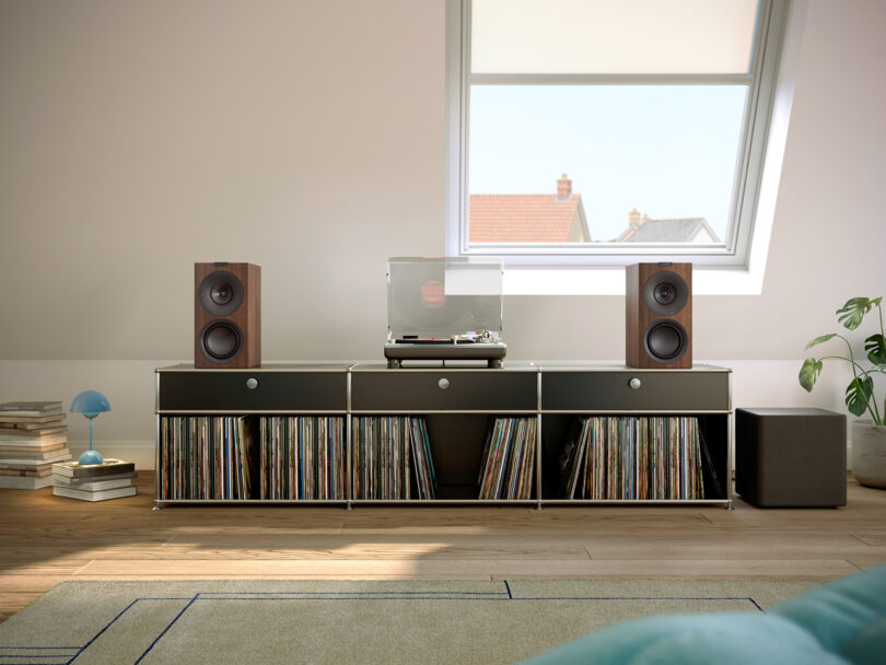 A vinyl record player with speakers is placed on a black storage unit filled with records. The unit is positioned near a window in a room with wooden flooring.