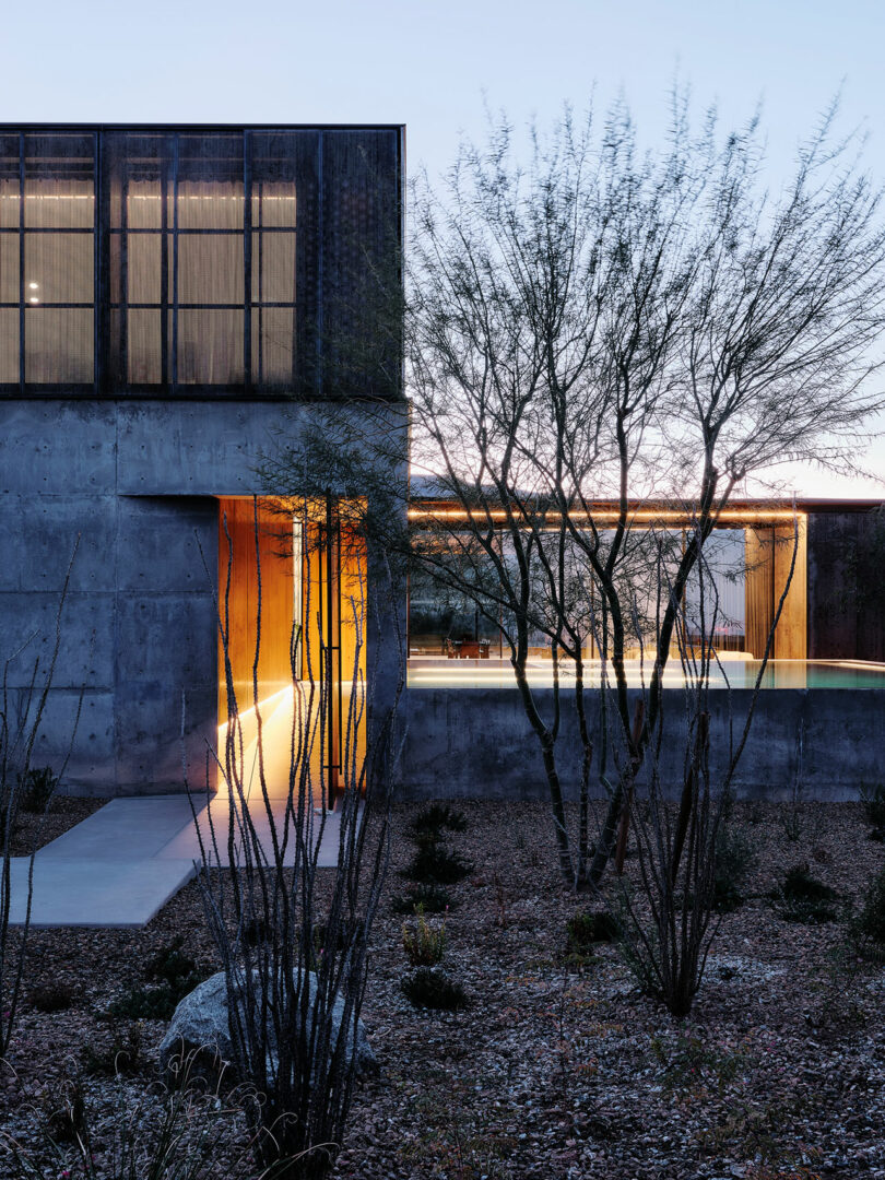 Modern concrete house at dusk with large windows, warm interior lighting, and sparse desert landscaping including bare trees and shrubs.